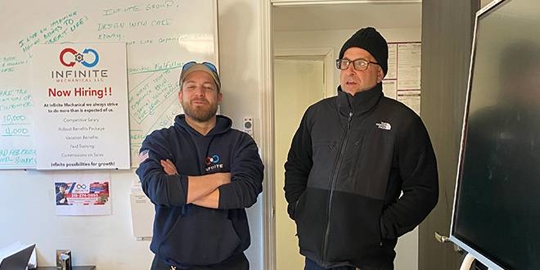Two men stand side-by-side in front of an office whiteboard.