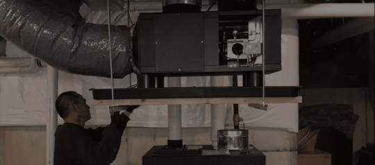 A person in a dark sweater is reaching up toward a black furnace in a basement.