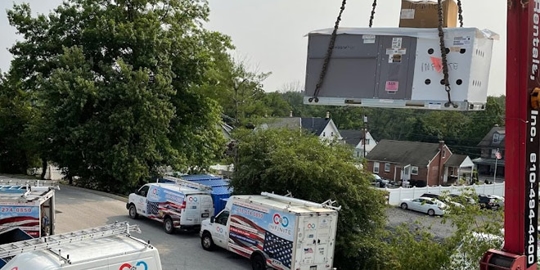 A crane lifts a new HVAC unit over service vans parked on the side of a building with patriotic branding